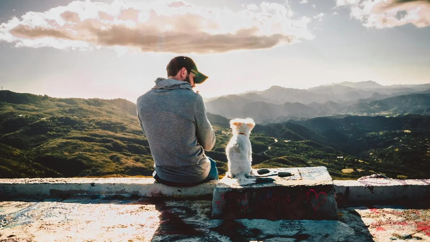 Mann sitzt mit einem Hund auf einer Mauer. Im Hintergrund sind Berge zu sehen.